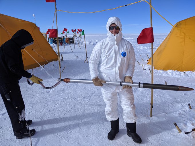 A man in a white sterile suite holds the Ice Diver (a long cylinder pointed at one end) horizontally in both hands. Ice Diver would be his height if stood upright. They stand on a field of white snow/ice with bright yellow tents in the background.