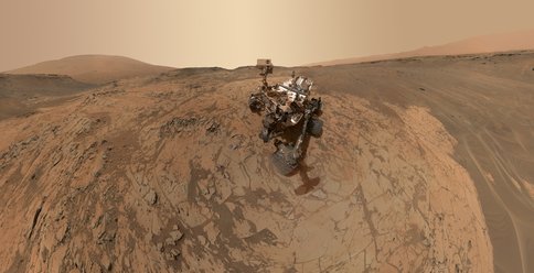 Figure 1. Self Portrait of Curiosity rover at "Mojave" drill site: This scene is a composite of dozens of images taken by the Mars Hand Lens Imager (MAHLI) at the end of the rover's robotic arm. The "Pahrump Hills" outcrop surrounds the rover, with Mt. Sharp visible in the back left, and the rim of Gale crater visible in the back right. 
