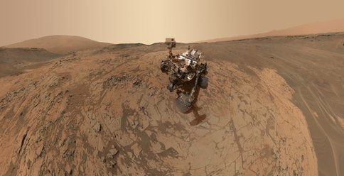 Figure 1. Self Portrait of Curiosity rover at "Mojave" drill site: This scene is a composite of dozens of images taken by the Mars Hand Lens Imager (MAHLI) at the end of the rover's robotic arm. The "Pahrump Hills" outcrop surrounds the rover, with Mt. Sharp visible in the back left, and the rim of Gale crater visible in the back right. 
