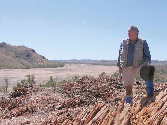 Jack farmer poses with one knee stepping up on a rock outcrop at the right of the frame. The background shows a dusty, desert landscape with hills and little brush.