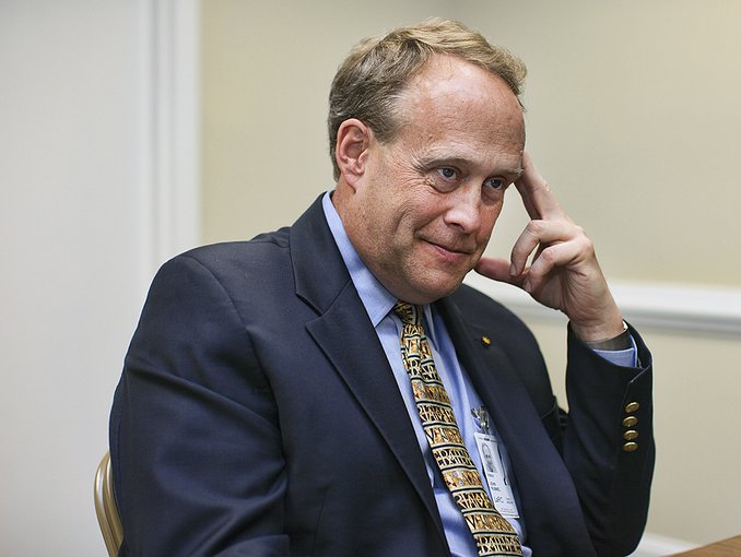 John Rummel sits at a table and is visible from the chest up. He smiles and looks off to the the right of the frame, holding his left hand up to his temple. He is wearing a blue suit with a gold tie.