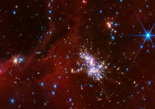 Image shows a dark background of reddish, cloud-like dust dashed against the black background of space. Bright points of light flare up in tints of blue, yellow and white at different sizes. A cluster of these points of light sits off center lower right.