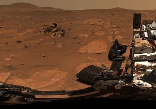 Wide image of the red, rocky, dusty surface of Mars. At the top of the page is an uneven horizon with hills and mountains. At the bottom of the frame parts of the rover can be seen.