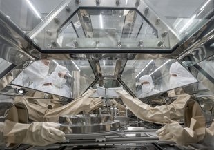 The view shows the inside of a sample handling chamber. It is glass. Outside the chamber four researchers can be seen in full 'bunny' suits. Their hands are in gloves built into the chamber so that they can handle a metal dish containing samples.
