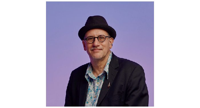A man with grey facial hair and round black glasses smiles softly at the camera, wearing a black fedora, black blazer, and blue floral shirt. He poses in front of a purple background.