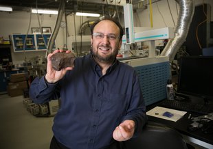 Jason leans on a counter in a lab full of equipment. He is wearing a long-sleeved dark blue button-up shirt. He has dark hair, a short beard, and glasses. Smiling broadly, he holds a palm-sized meteorite fragment up for the camera in his right hand.