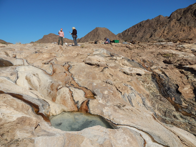 Alta Howells (left), Alysia Cox (standing right) and James Leong (seated) take a gas sample for H2 analysis, review field notes and analyze aqueous chemistry on a spectrophotometer in Oman.