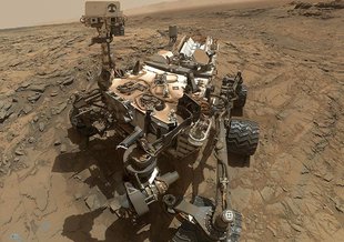 In this image, composed of dozens of photographs, the Mars Curiosity rover is pictured next to a drill hole it has made in the martian surface, seen in the lower left-hand corner.