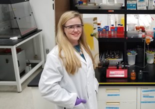 Barge stands in front of a lab bench with equipment on it. The shelves above the lab bench are full of test tubes and other supplies. Barge has long, blond hair past her shoulders. She wears safety glasses, a white lab coat, and purple lab gloves.