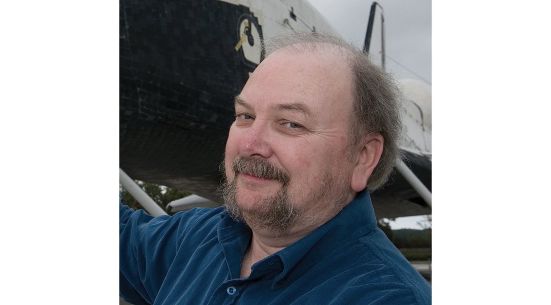 A close up of Nicholson's smiling face. He is wearing a blue button-up shirt. He has a goatee and thin, light brown, wispy hair. Part of a Space Shuttle are visible in the background.
