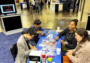 Four people sit at a table in the conference center intently looking at pieces of a board game scattered across the top of the table.