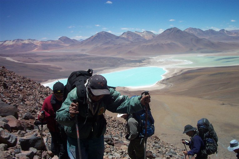 The team is in the foreground, hunched under their backpacks and climbing toward the camera up a mountain. In the distance, the bright blue water of a lake can be seen among the rocky, desert landscape.
