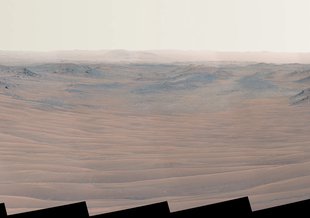 This long horizontal image shows the reddish landscape of Mars. At the left and right edges are rocky banks. Central to the image is the trough of a dry riverbed filled with sandier material.