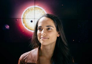 Portrait from the shoulders up. Natasha wears a brown leather jacket and her dark eyes looks off to the left. She is smiling, lips together. Her long, straight, dark hair drapes across her shoulder. Backdrop of dark space with planets orbiting a star.