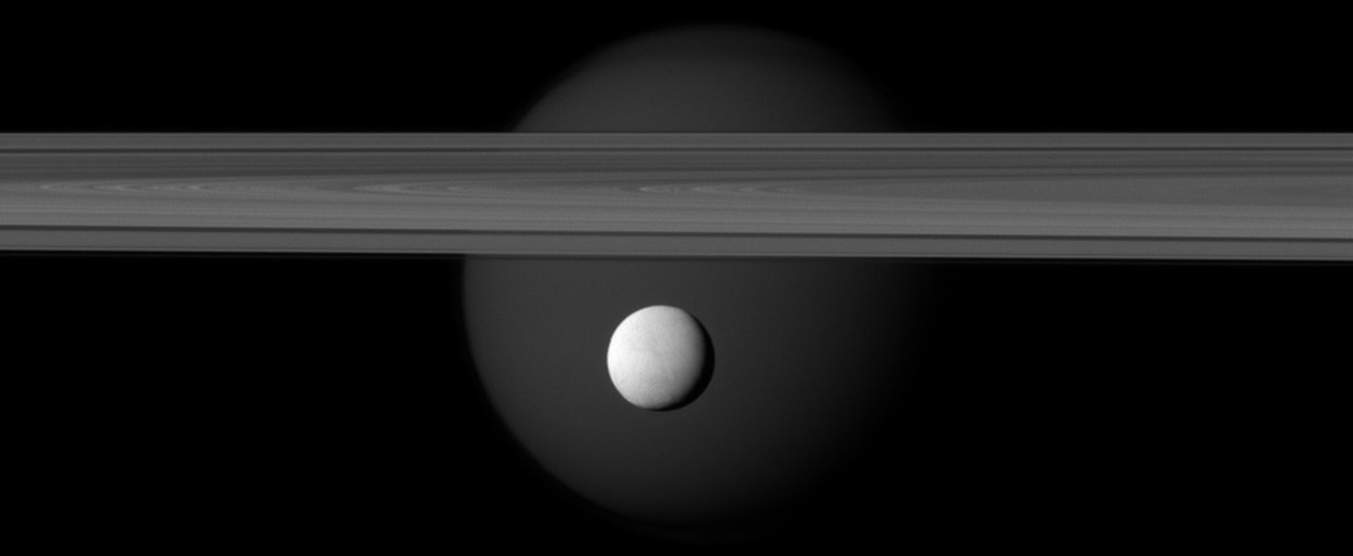 A brightly reflective Enceladus appears before Saturn's rings, while the planet's larger moon Titan looms in the distance. Jets of water ice and vapor emanating from the south pole of Enceladus hint at a subsurface ocean on the tiny moon.