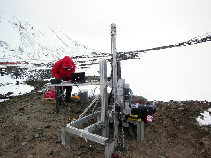 Icebreaker is in the foreground. The sky is a hazy white. A researcher in a bright red jacket hunches over a portable table with computer equipment in the background. Snow is prominent on the ground and the mountain face in the distance left of frame.