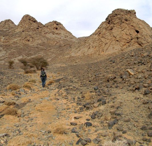 A woman walks a rocky path through a desert. Boulders and the dry, dusty mud mounds are seen in the near distance.