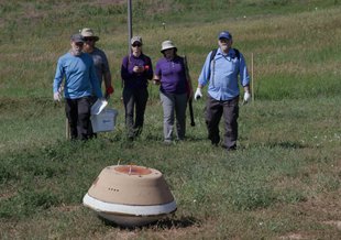 A group of researchers walk through a grass-covered landscape toward a capsule in the foreground. The landscape is hilly so that no sky is visible in the background.