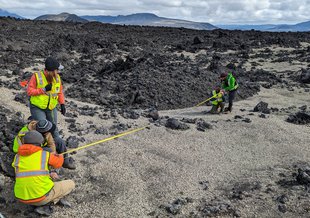Three researchers in high visibility gear are in the foreground left facing away from the camera. Across the terrain of volcanic rock and pebbles, two researchers can be seen in the distance. Two groups stretch a measuring tape out between them.