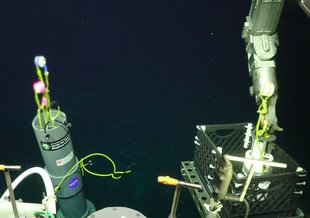 Against an almost black background of deep seawater, the arm of the ROV Jason vehicle is placing samples in a basket. Parts of the top of Jason are visible, including a pipe with a NASA logo.