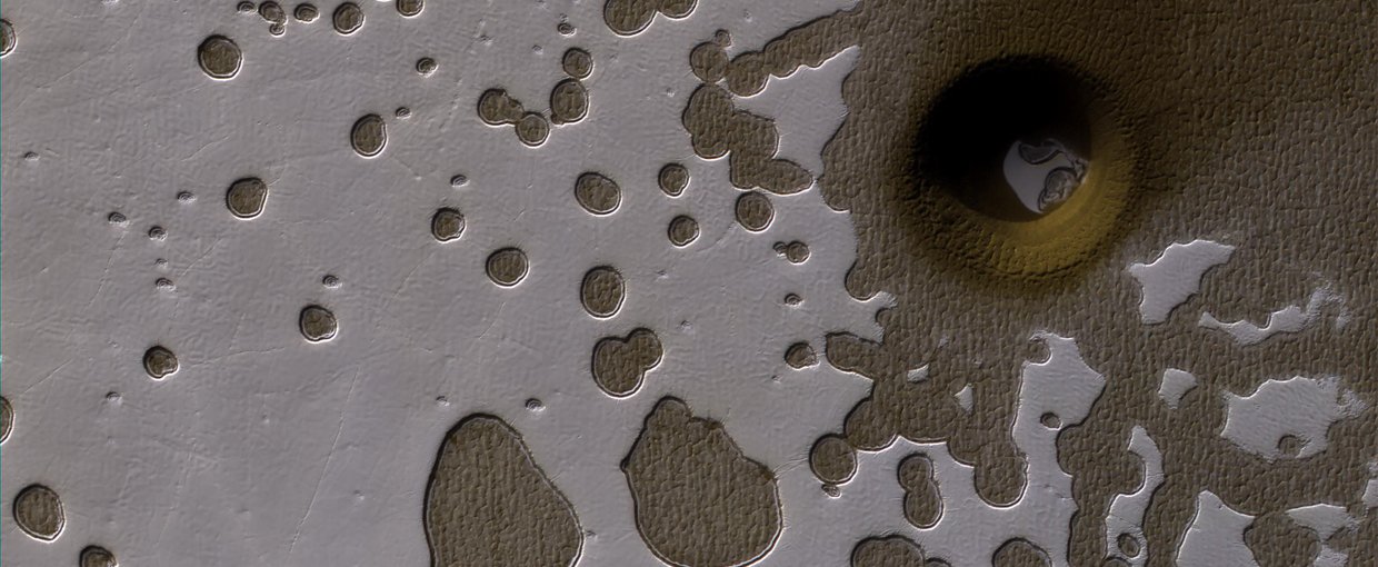 MRO image in the Southern hemisphere of Mars. There are shallow pits in the bright residual cap of carbon dioxide ice and a circular formation that penetrates through the ice and dust. This might be an impact crater or it could be a collapse pit.