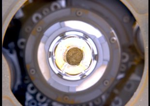The image is a close up looking straight through the inside of a sample tube housed in circular metal components of the rover with visible screws holding the instrument together. Light shines down inside the tube filled with a small circle of rock.