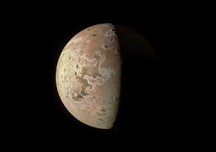 The right half of the sphere of Io is visible against an inky black backdrop. The other half of the moon is shrouded in darkness. What can be seen of Io is a pock-marked surface of craters and mountains that appear in shades of yellow, orange and tan.