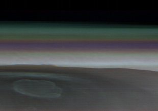 A very wide landscape image of Olympus Mons from orbit. The caldera of the volcano is in the center. The rest of the planet appears shrouded in thin cloud cover. A thin streak of colors from purple to green runs along the horizon as the atmosphere catches
