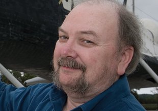 A close up of Nicholson's smiling face. He is wearing a blue button-up shirt. He has a goatee and thin, light brown, wispy hair. Part of a Space Shuttle are visible in the background.