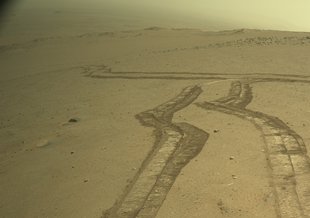 This image shows two lines of tracks left by the rover as it traversed a section of Mars. The tracks twist away into the distance, disappearing over a ridge. In the light, the image appears to have a yellowish tint with a dusty dissolve over the distance.