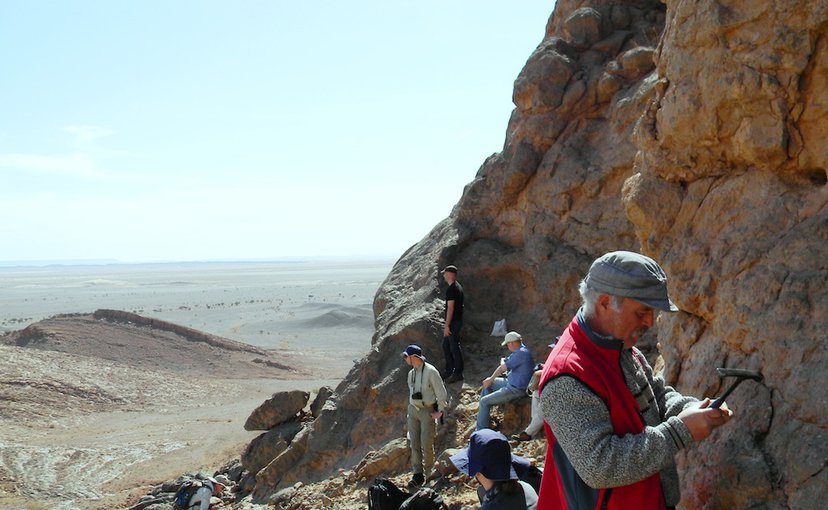 A group of people hug against a sheer wall of the mountainside. In the foreground a man with a mustache uses a rock hammer to tap at the wall. The desert spreads out in the distance.