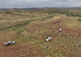 An overhead photograph of trucks driving on a dirt road through scrub-covered hills in Australia under an overcast sky.
