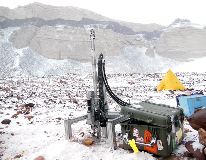 The Icebreaker drill stands in the foreground on a rocky, snow-covered terrain. Mountain walls are visible through haze in the background. A yellow pyramid tent is also visible. Next to the drill are heavy-duty plastic boxes containing field equipment.