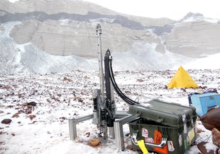 The Icebreaker drill stands in the foreground on a rocky, snow-covered terrain. Mountain walls are visible through haze in the background. A yellow pyramid tent is also visible. Next to the drill are heavy-duty plastic boxes containing field equipment.