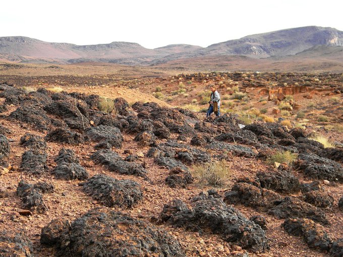 In the distance are dry, rocky mountains. A figure walks through the middle distance on sloping hills of rock and sand. In the foreground are low, dark, rock-like mounds that poke up out of the desert floor.