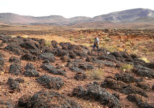 In the distance are dry, rocky mountains. A figure walks through the middle distance on sloping hills of rock and sand. In the foreground are low, dark, rock-like mounds that poke up out of the desert floor.