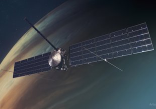 The wide reach of Clipper's solar panels fills most of the width of the landscape image. The spacecraft is centered in frame with Jupiter in the background. Jupiter appears muted and hazy, with Clipper in focus in the foreground.