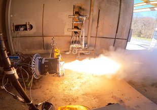 A rocket at the left of the frame fires in a garage facility. The flame from the rocket shoots out to the right and out of the door of the garage.