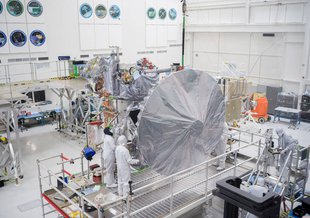 A group of engineers in bunny suits examine the Europa Clipper mission in a clean room at NASA JPL. The spacecraft is in the center wrapped in silver material. The photo is from a high angle looking down at the spacecraft and engineers.