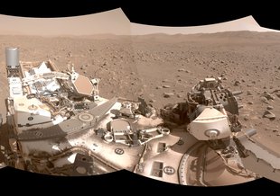 A wide angle shot from Perseverance of the sandy, rocky surface of Mars. Parts of the rover can be seen in the center foreground. Mountains are seen in the distance.