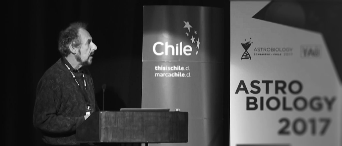A black and white photo of Andrew standing behind a podium and speaking. The photo is slightly from the side and banners for the conference are seen in the background.