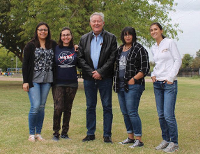 Jack Farmer stands in the middle of a group of students. They are outside on a clear day, standing in the grass with leafy trees behind them.