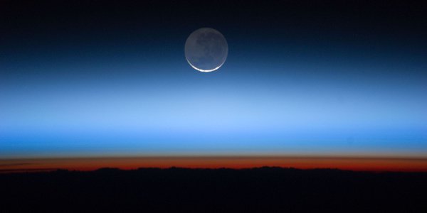 Image showing an orange glow in the lowest layer of the Earth's atmosphere. This glow comes from the Sun, which is just below the horizon. The Moon glows softly at the top center of the image as the orange light fades to blue and then the black of space.