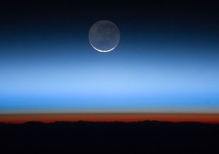 Image showing an orange glow in the lowest layer of the Earth's atmosphere. This glow comes from the Sun, which is just below the horizon. The Moon glows softly at the top center of the image as the orange light fades to blue and then the black of space.