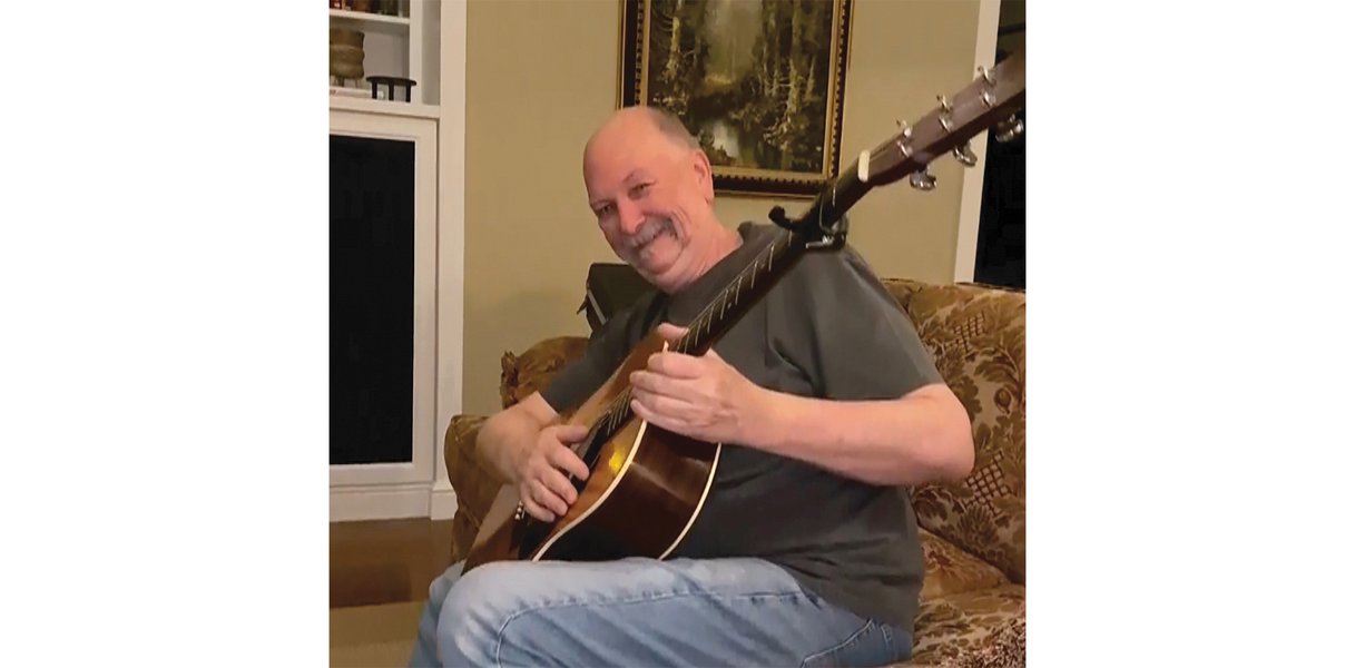 Wayne in jeans and a dark t-shirt sits on a brown floral-patterned couch holding an acoustic guitar. The image is slightly side on, with Wayne laughing and leaning away from the camera.