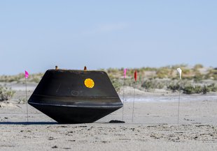 The blackened, somewhat cone-shaped capsule sits on it's "nose" (left of frame, side-on view) on sandy ground with scrub grass in the background. Small flags are stuck in the sand around the capsule.