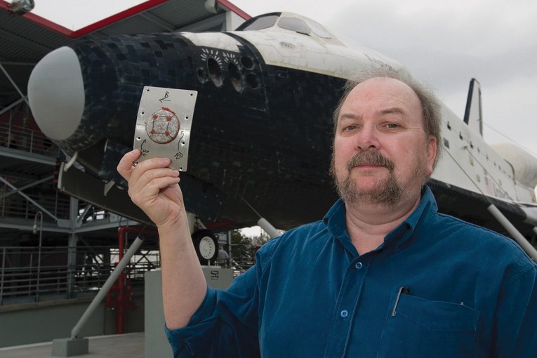 Nicholson stands to the left of frame in a blue button up shirt with sleeves rolled to the elbow. He is holding up a rectangular metal panel. In the middle of the panel is a circle of simulated meteorite material. Parked behind him is a Space Shuttle.