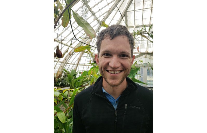 Noah smiles in the foreground wearing a black half-zip jumper. He is in a greenhouse with plants visible in the background along with the windowed roof of the glasshouse.