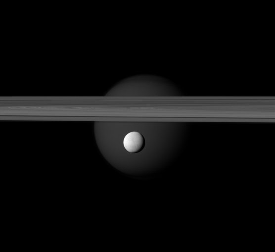 A brightly reflective Enceladus appears before Saturn's rings, while the planet's larger moon Titan looms in the distance. Jets of water ice and vapor emanating from the south pole of Enceladus hint at a subsurface ocean on the tiny moon.