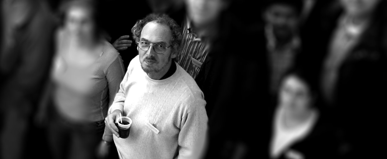 Black and white photo of Andrew in a crowd of people at a conference. The angle of the camera is from above. Andrew looks into the camera wearing a white sweater and holding a cup of coffee. A focal blur means only he is in focus.